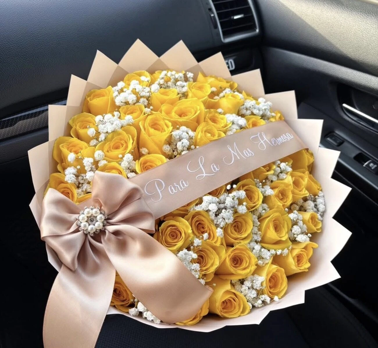 Flores amarillas con forma de rosa, pequeñas flores blancas, envoltura clara y un gran lazo rosa pálido.