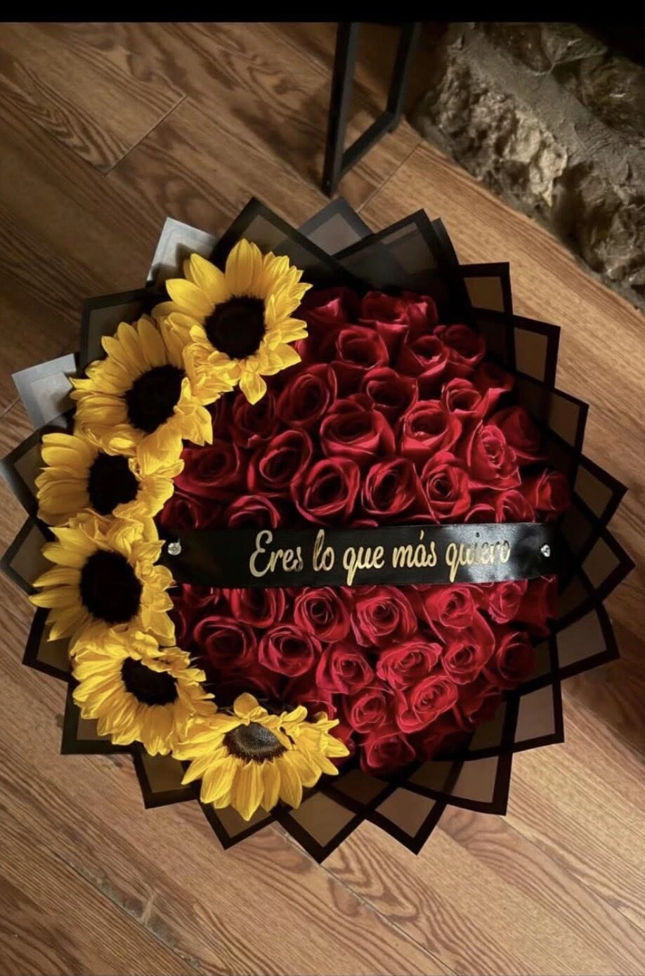 Flores rojas y girasoles dispuestos en círculo con envoltura oscura y una cinta central.