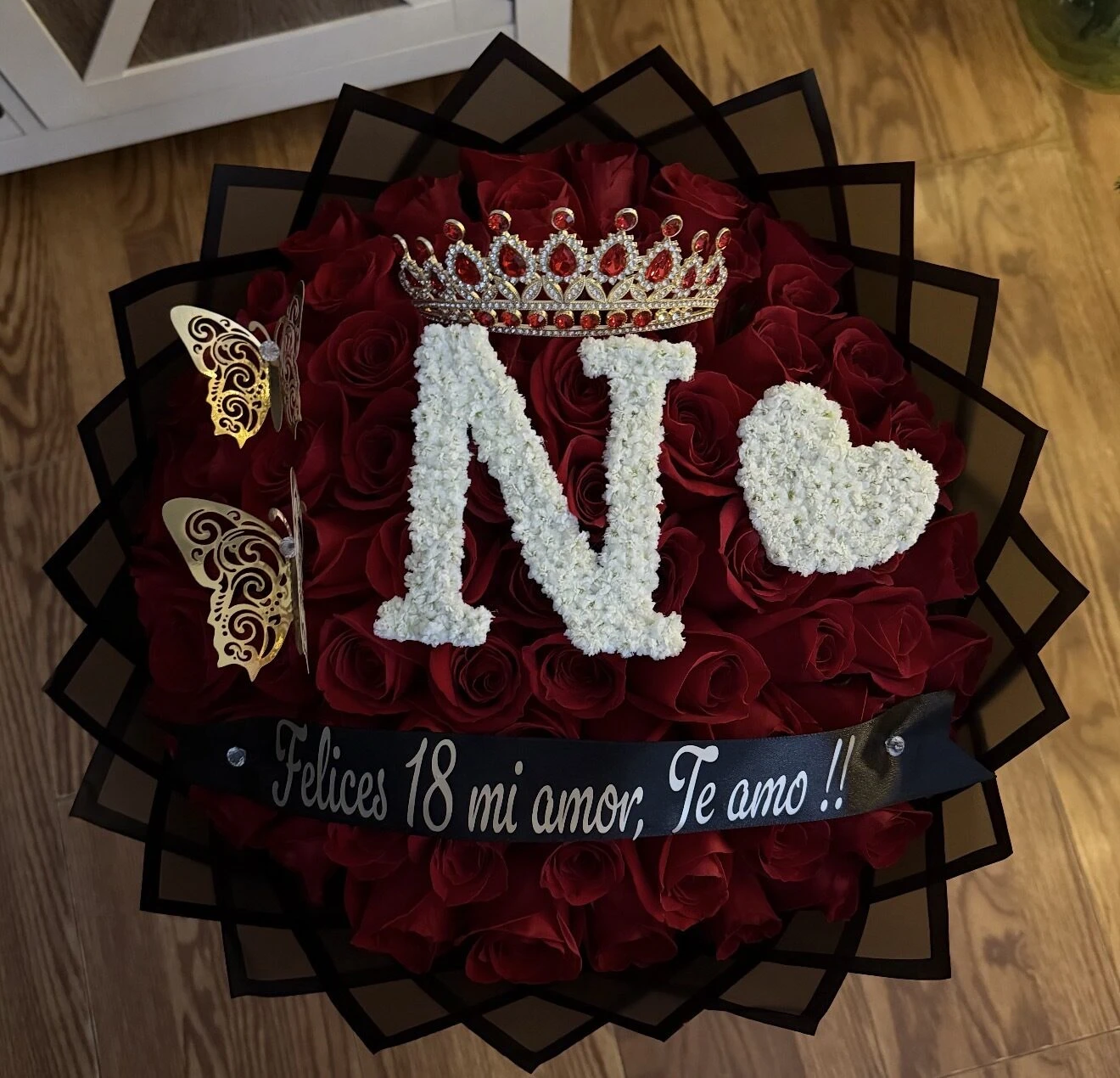 Arreglo floral rojo y negro con una letra blanca grande y un detalle de corazón.