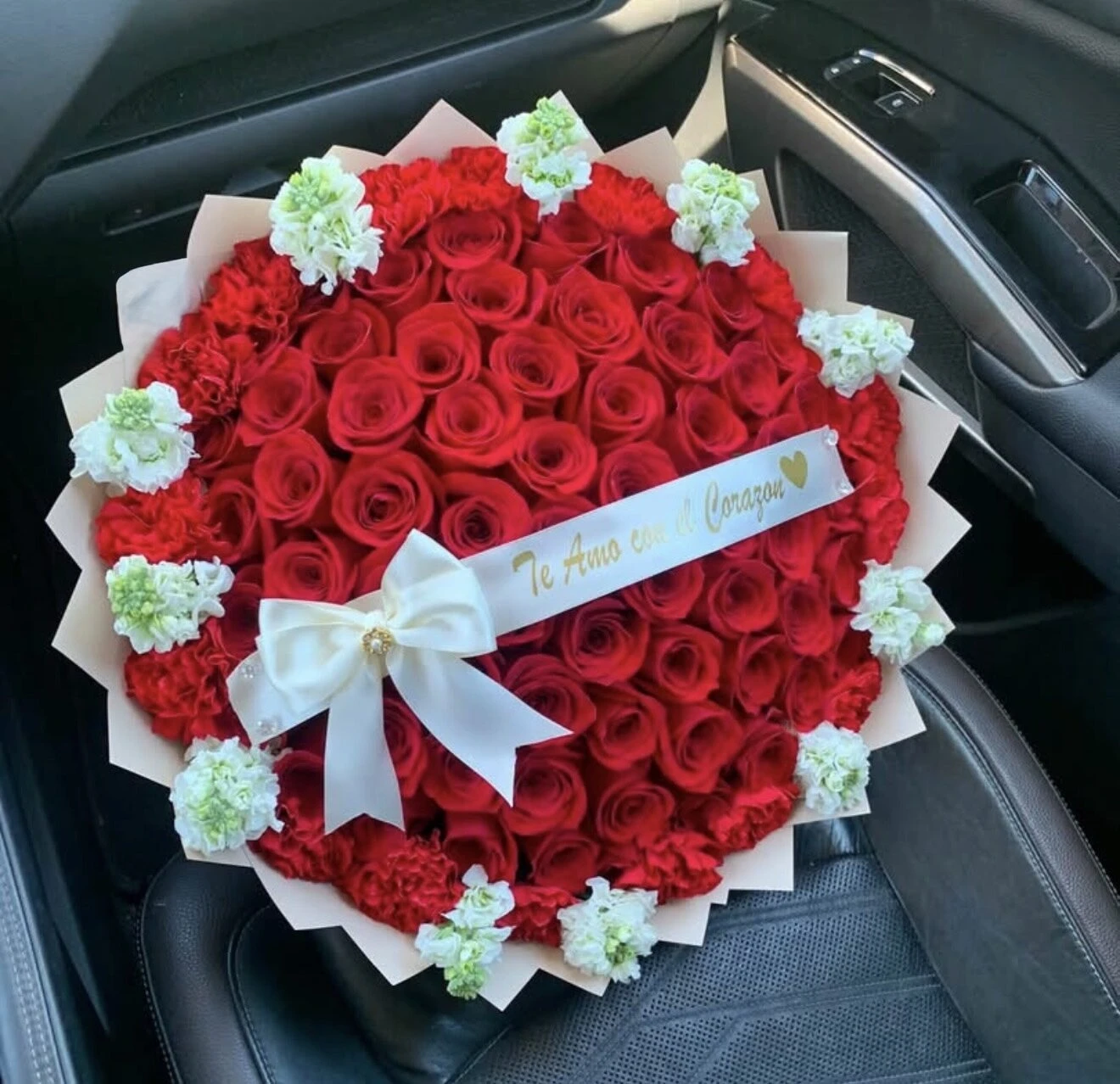 Arreglo floral rojo redondo con flores blancas y un lazo claro, fotografiado en un auto.